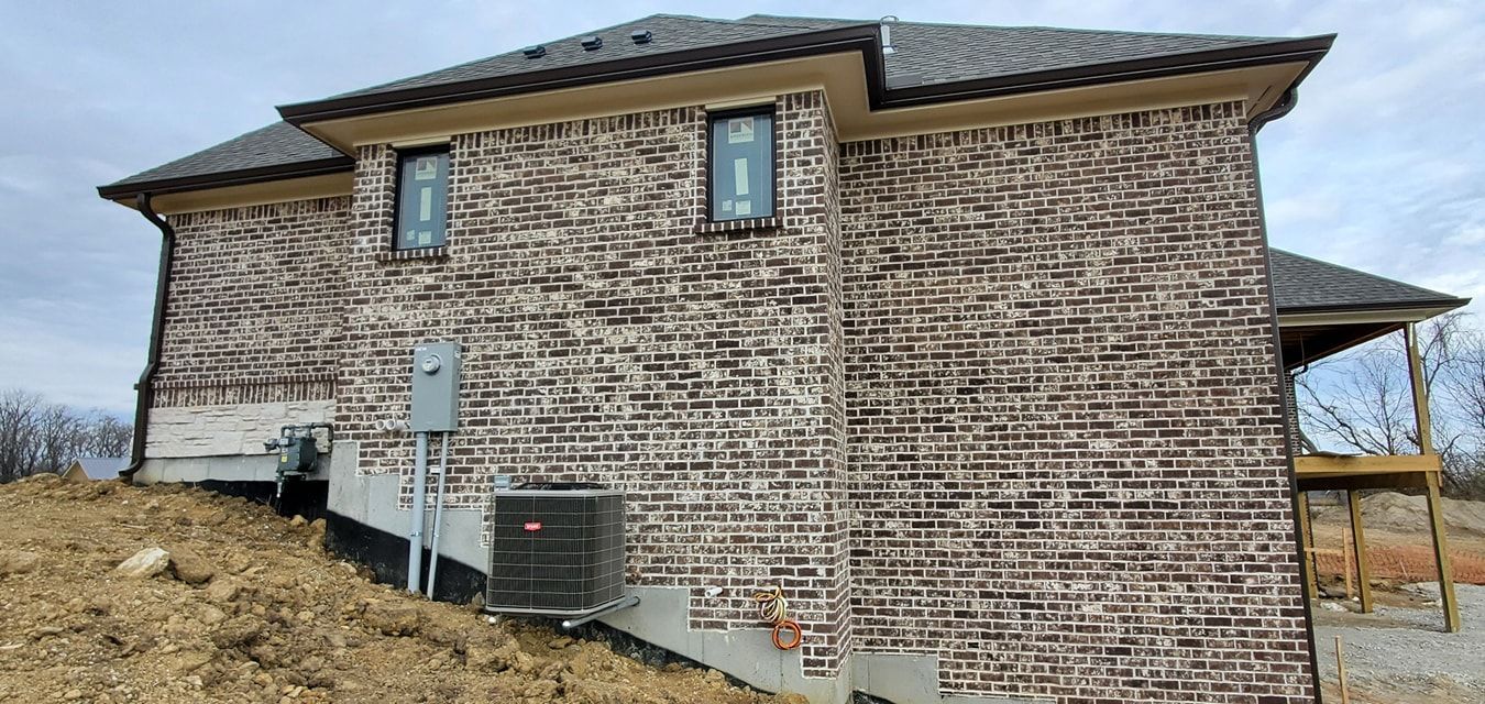 The rear exterior view of a new brick house with an air conditioning unit and exposed foundation on a dirt lot.