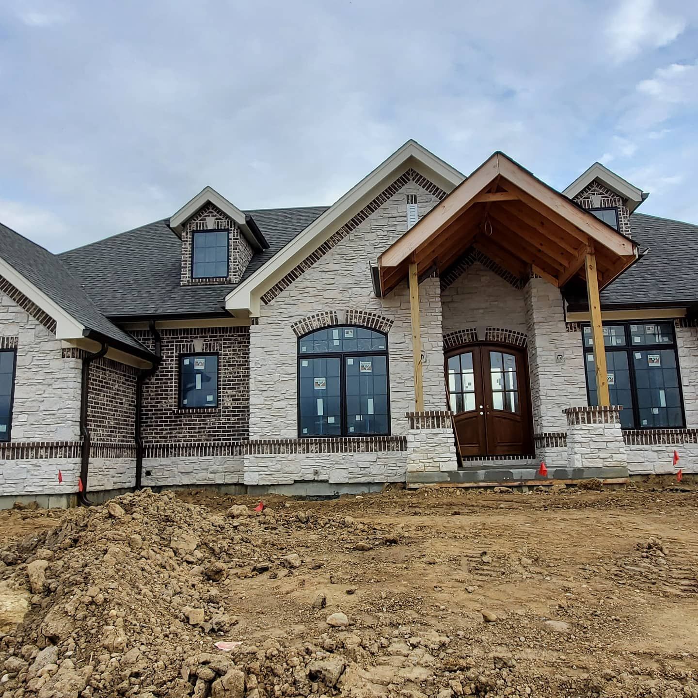 A newly constructed, light-toned stone house with a dark gabled roof, wooden front porch, and unfinished dirt yard.