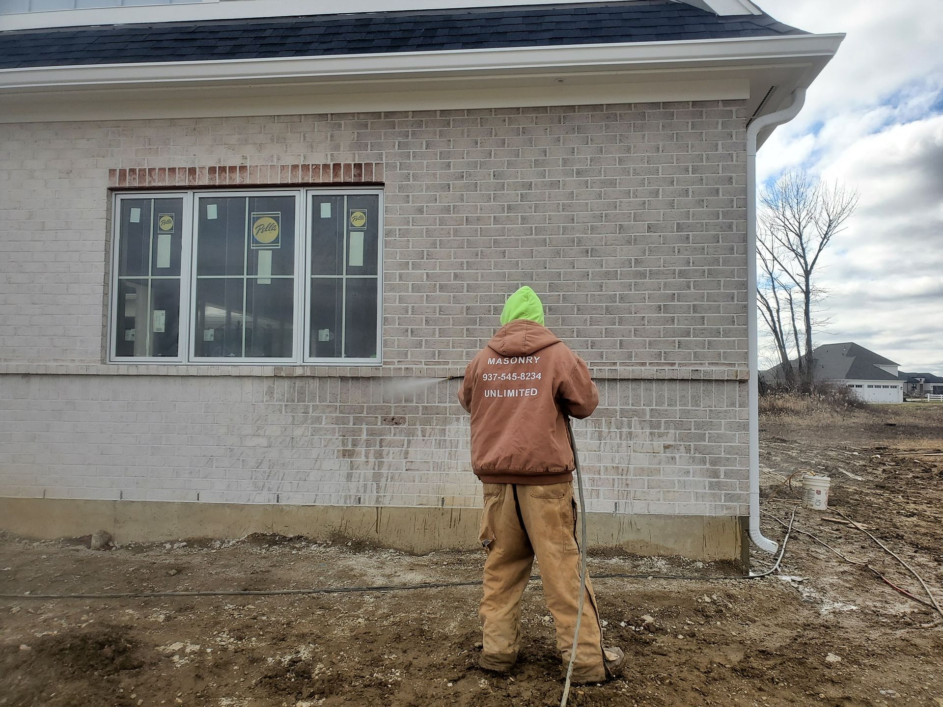 A worker in a brown jacket and bright green hat sprays a light-colored brick wall of a house under construction.
