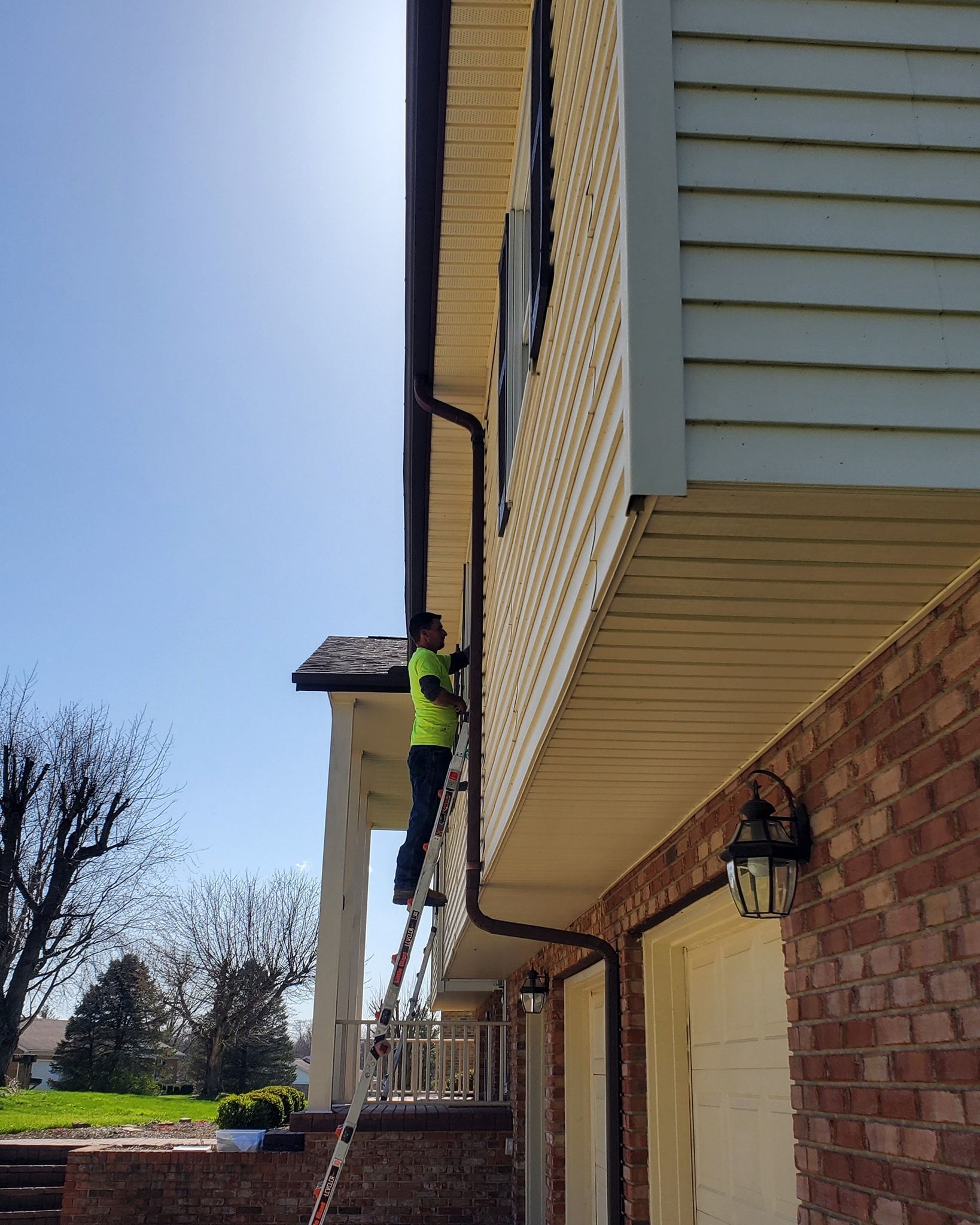 A worker in a bright yellow shirt climbs a ladder to perform maintenance on the siding and gutters of a brick house.