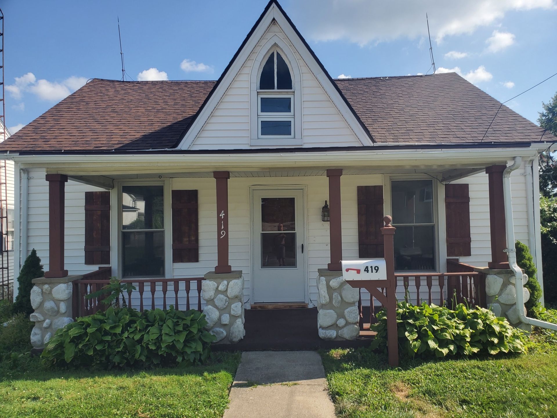 A white cottage with a front porch, brown shingled roof, arched attic window, and river rock porch pillars.