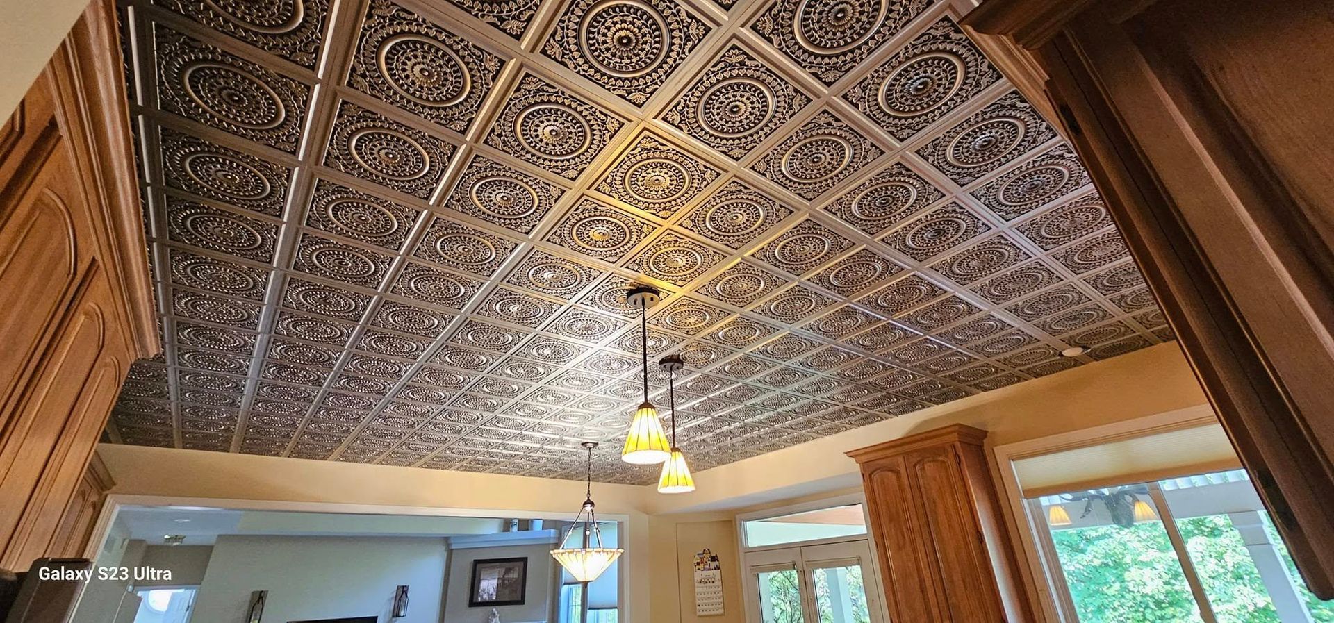 A low-angle view of a kitchen ceiling featuring intricate, dark-patterned metal tiles and two hanging pendant lights.
