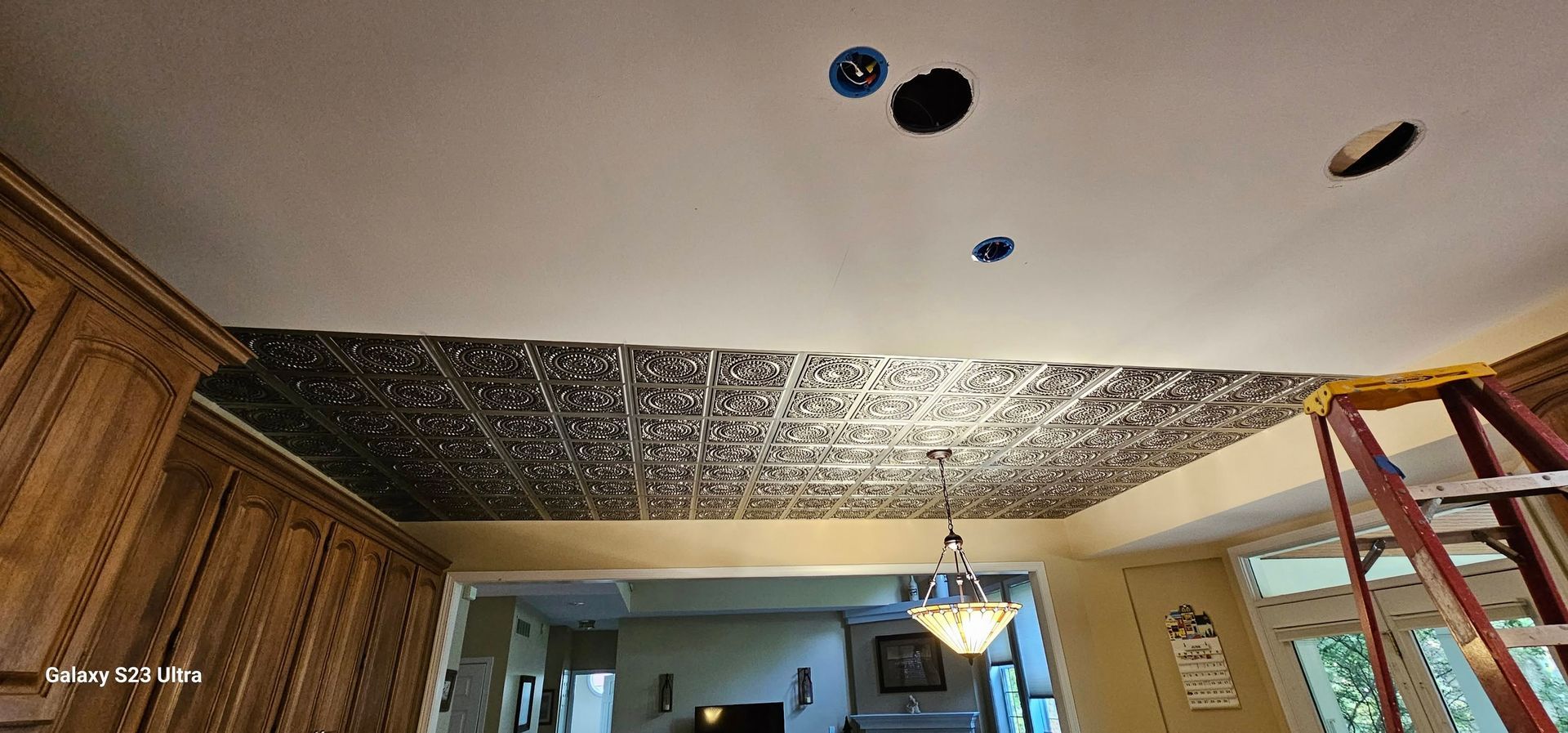 A partially installed metallic-style decorative ceiling tile panel in a room with a ladder, cabinets, and light fixtures.