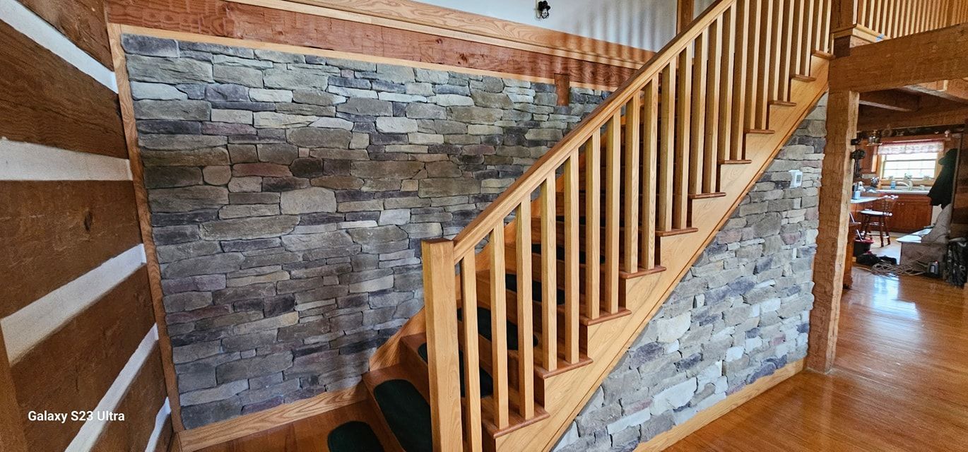 A wooden staircase with balusters rises beside a textured, multi-colored stone wall in a log cabin interior.