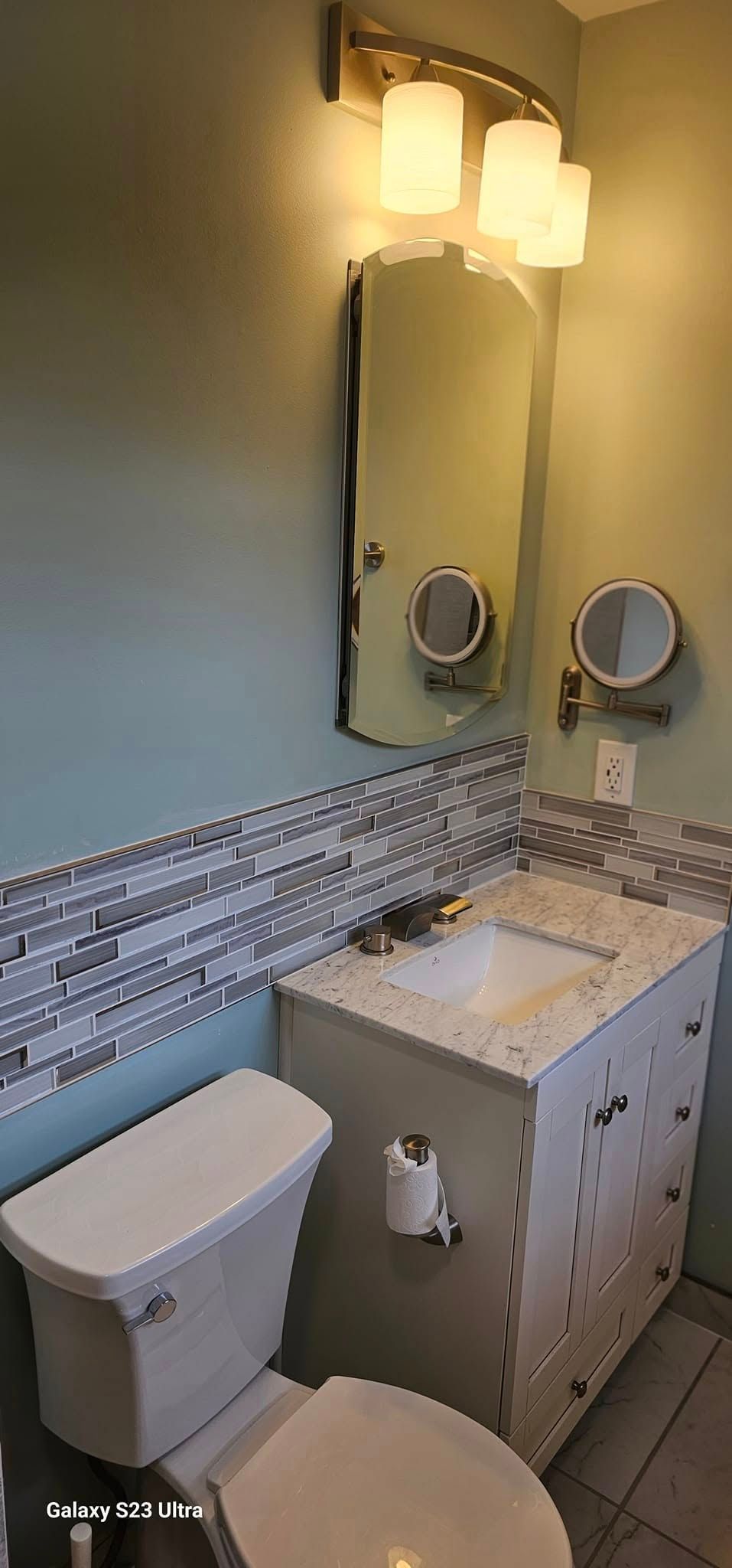 A bathroom vanity with a white sink, grey cabinet, decorative backsplash, a mirror, and a wall-mounted magnifying mirror.