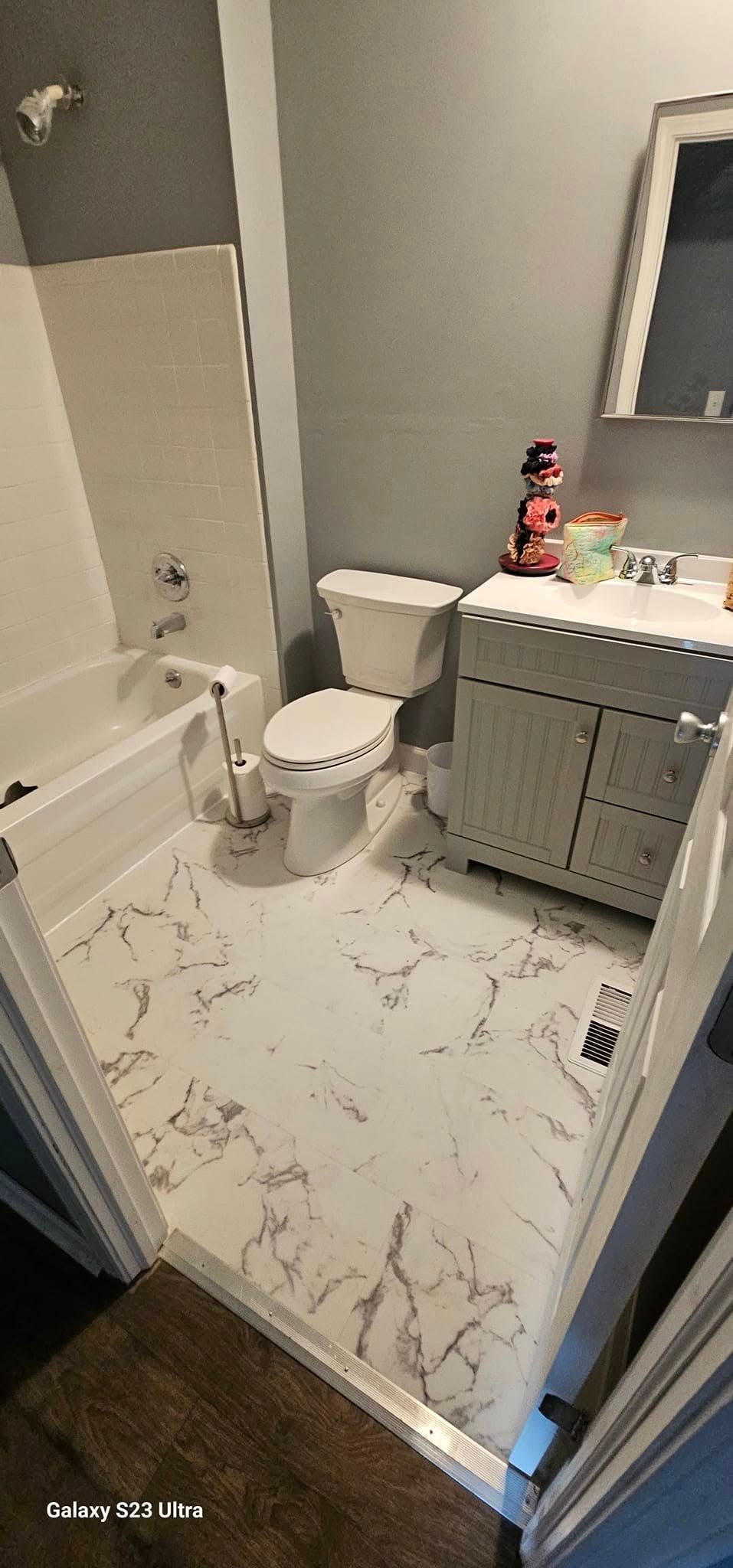 A small bathroom with a white vanity, toilet, and bathtub, featuring light-colored, marbled floor tiles.