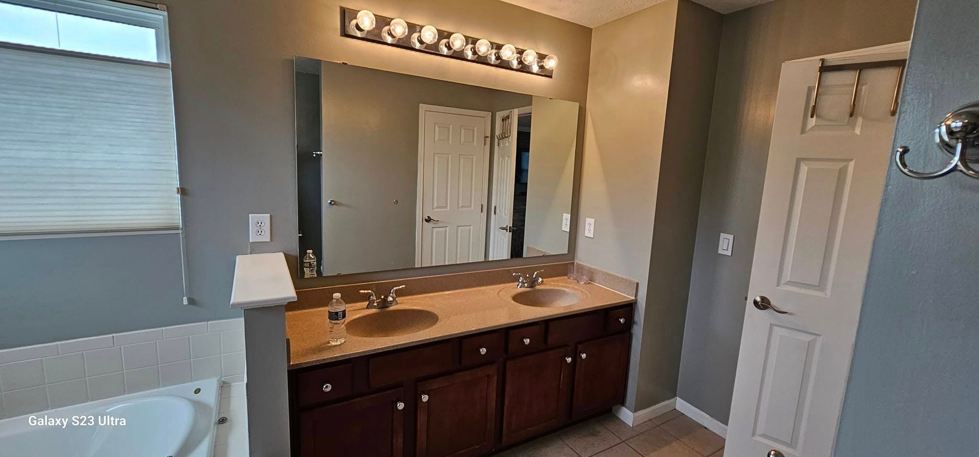 A bathroom with a double-sink vanity featuring dark wood cabinets, a large mirror, vanity lighting, and a white bathtub.