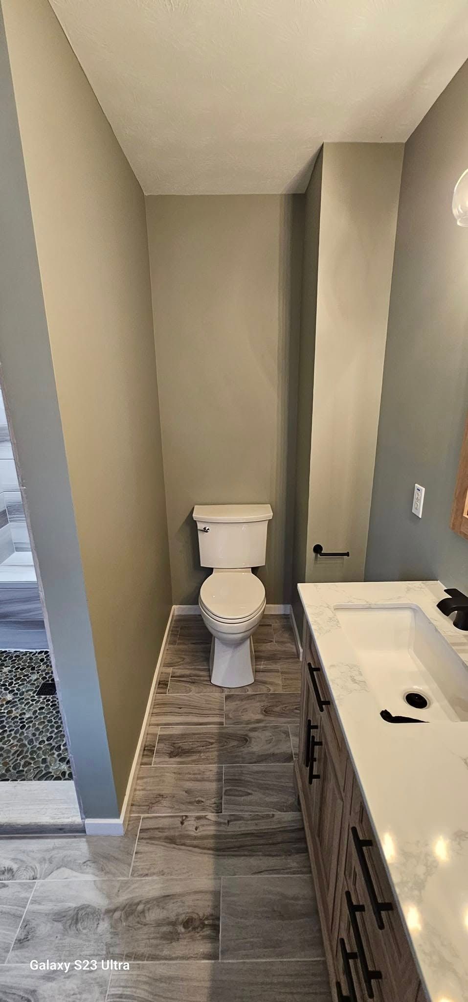 A modern bathroom featuring a white toilet between a taupe wall and a vanity with a white countertop and wood cabinets.