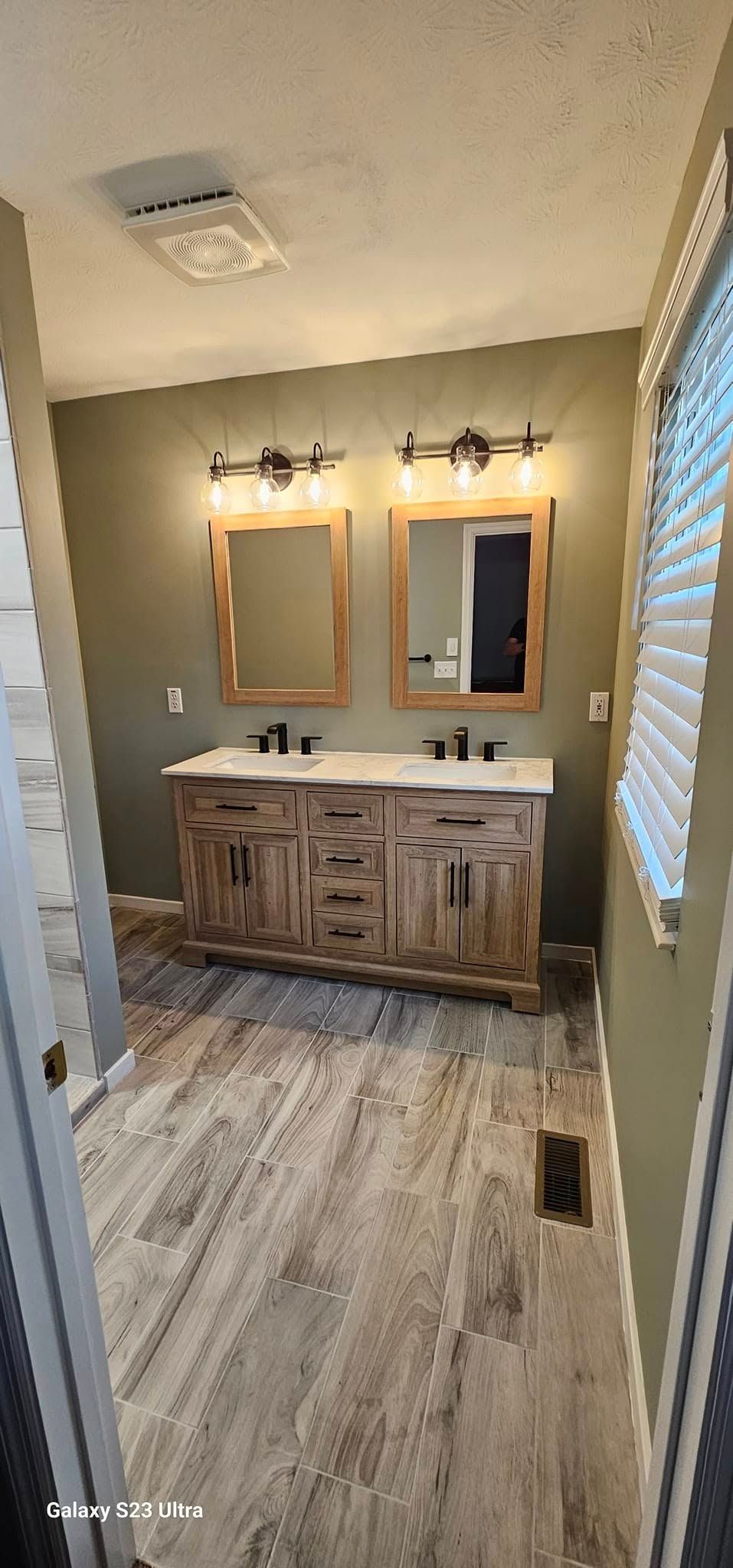 A rustic wooden double vanity with black hardware and matching mirrors against an olive wall in a bright bathroom.
