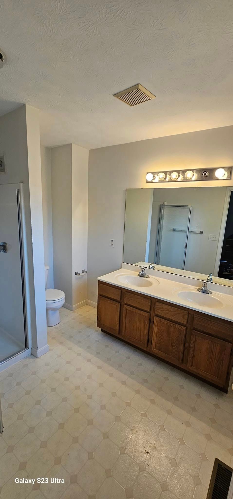 Bathroom with a wooden double vanity, white countertop, large mirror, and toilet tucked in a corner near a shower stall.