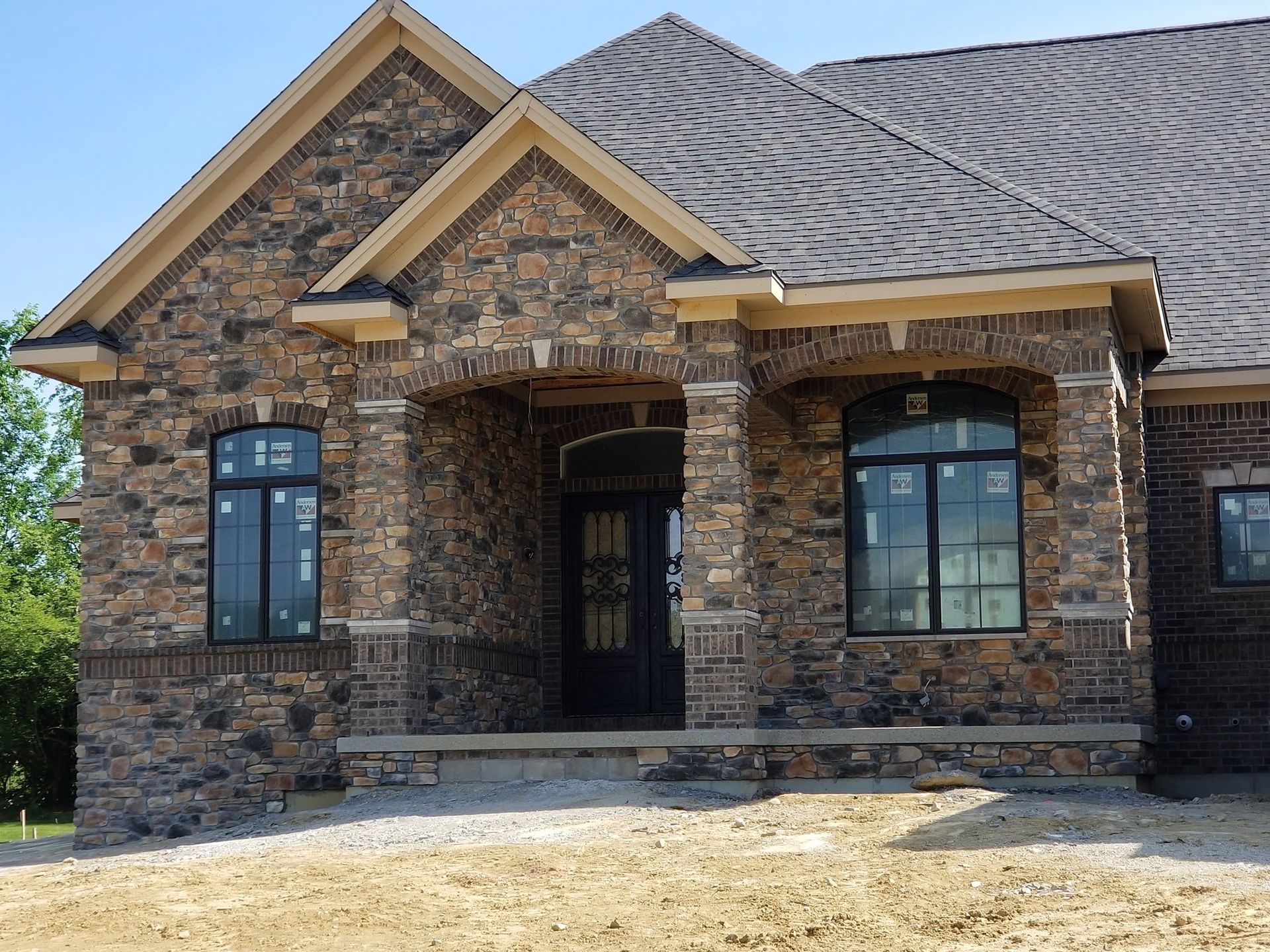 Single-story house with dark stone masonry, arched window frames, and a dark front door under construction.