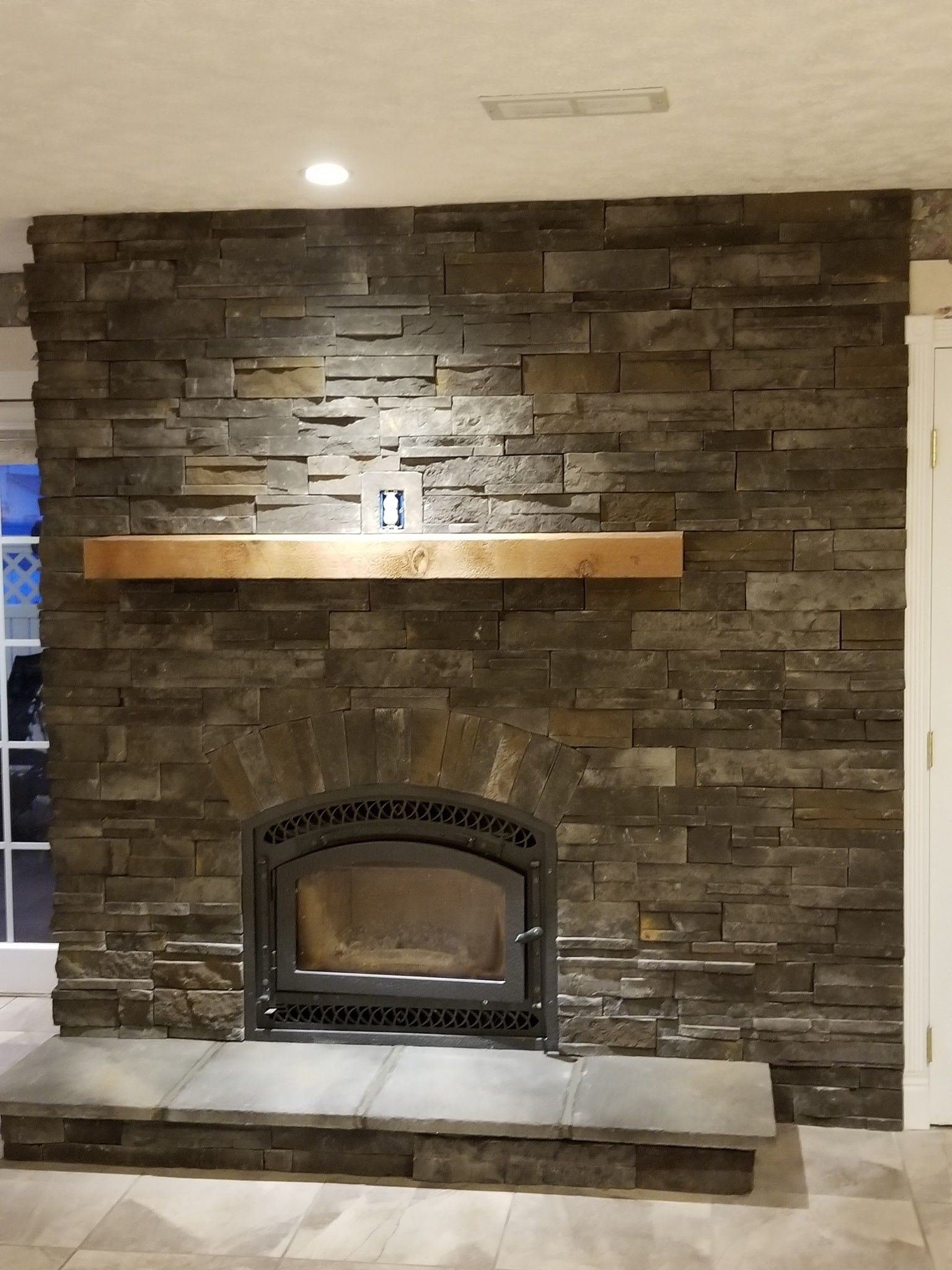 A dark-toned stacked stone fireplace with a thick natural wood mantel and a decorative black arched firebox.