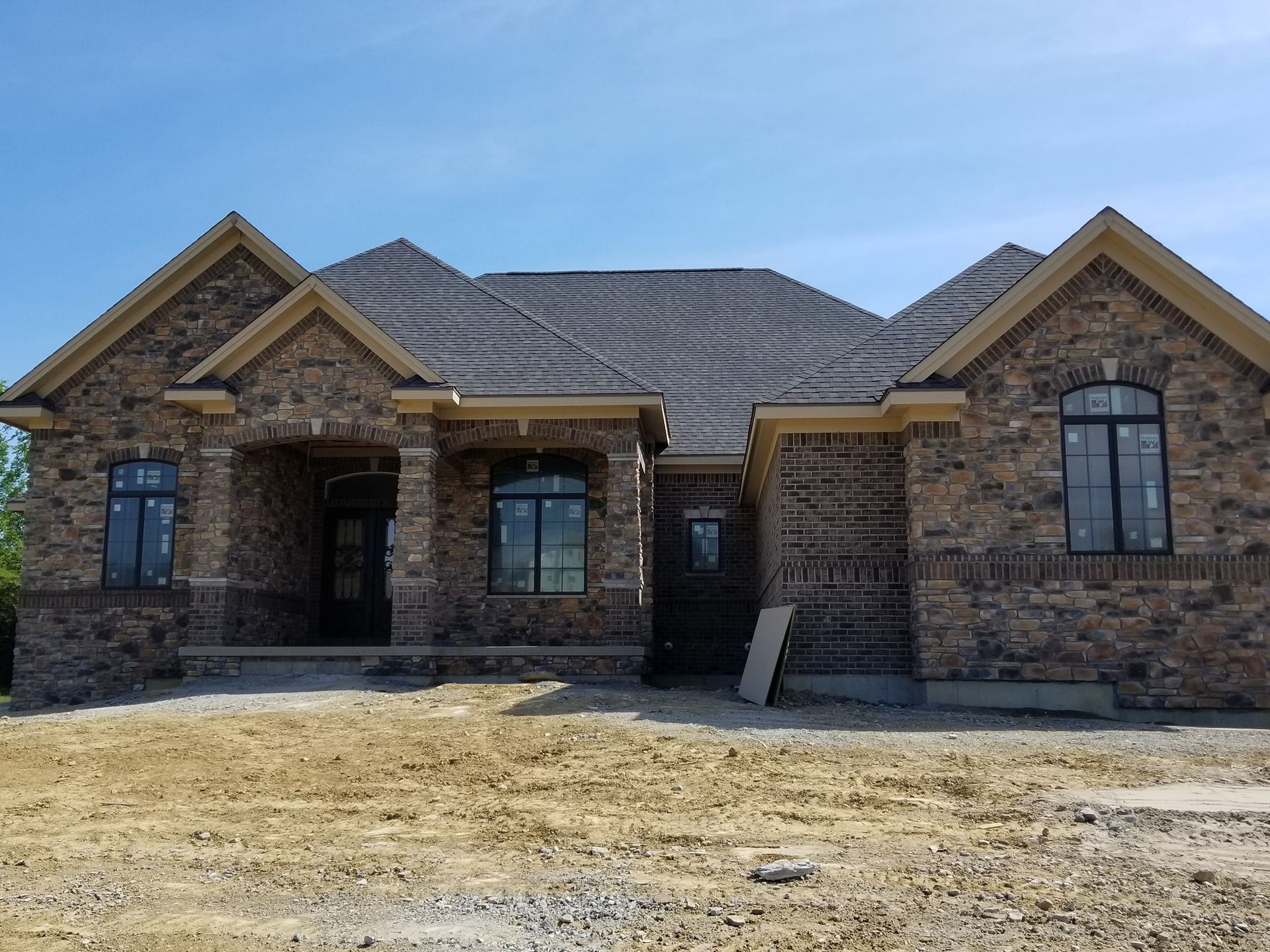 A newly constructed single-story house with a stone facade, arched windows, and a dark shingled roof under a blue sky.