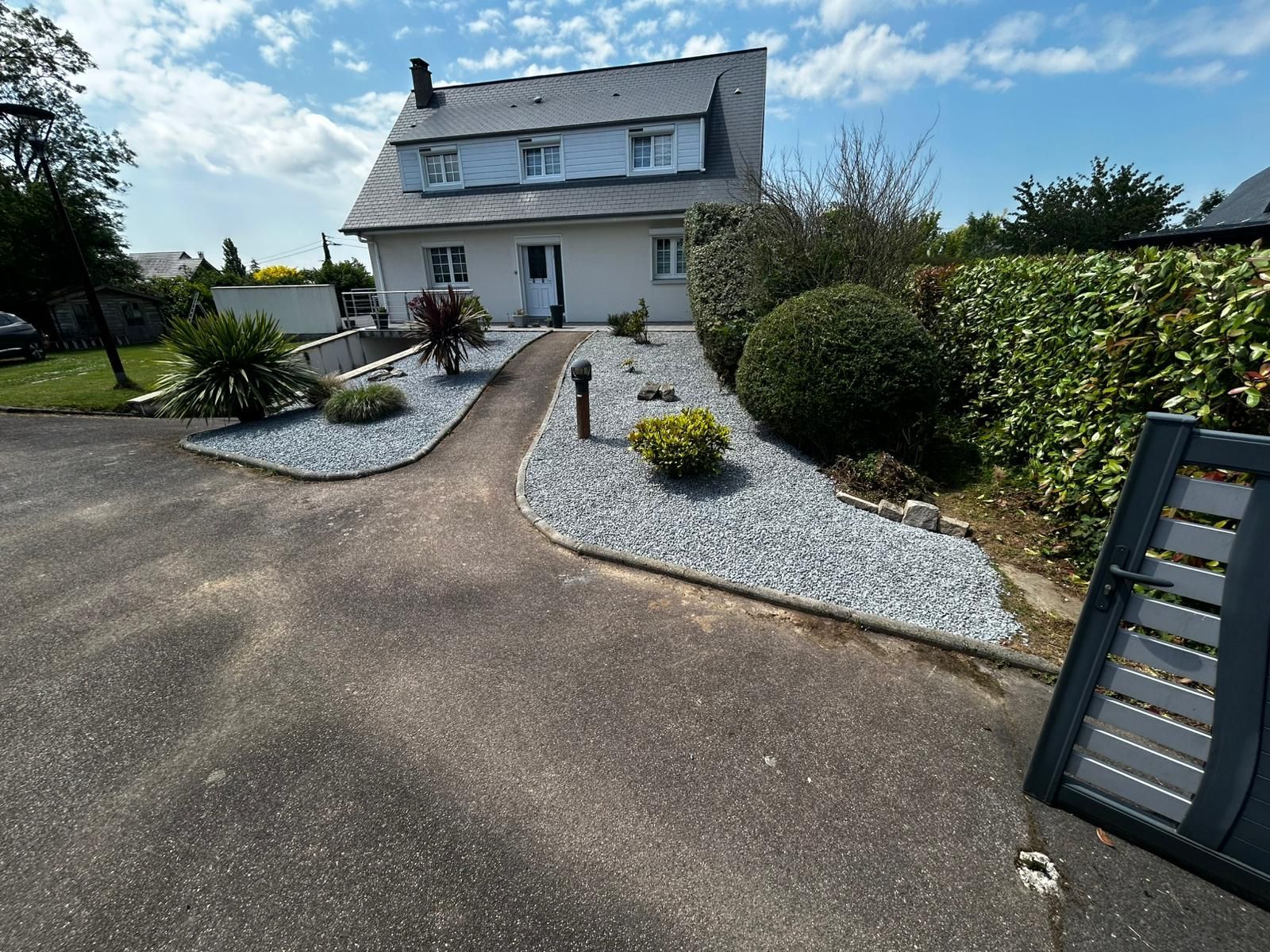 Une maison blanche au toit gris possède un chemin d'accès au jardin recouvert de pierres, entouré d'arbustes
