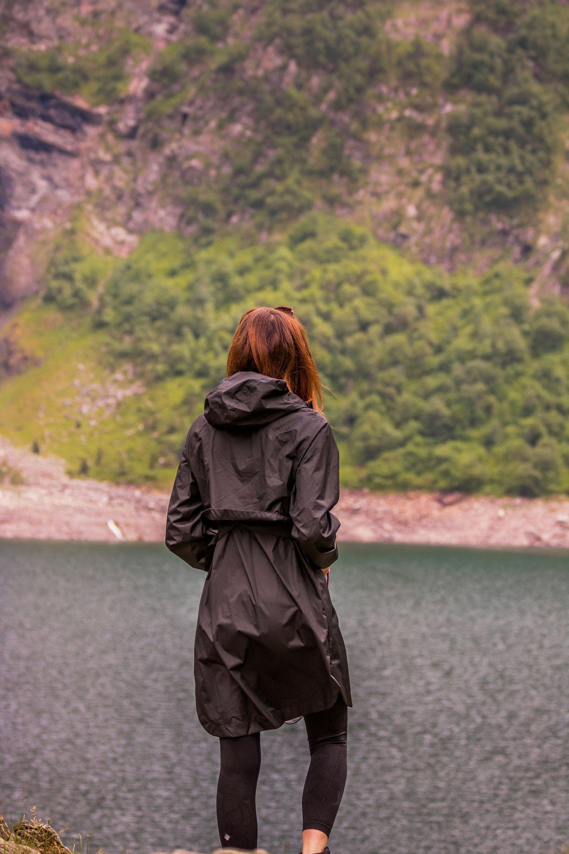Une personne vêtue d'un manteau noir à capuche se tient face à un lac calme, avec une montagne luxuriante et boisée