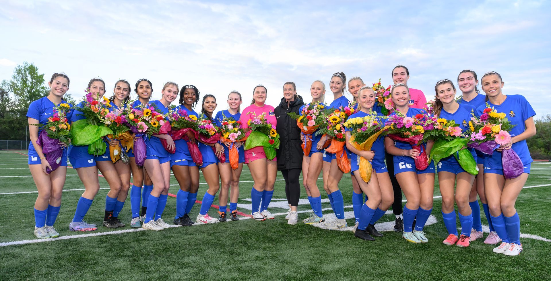 Senior players in tiaras and with flowers for Senior Night. head Coach in center with players.