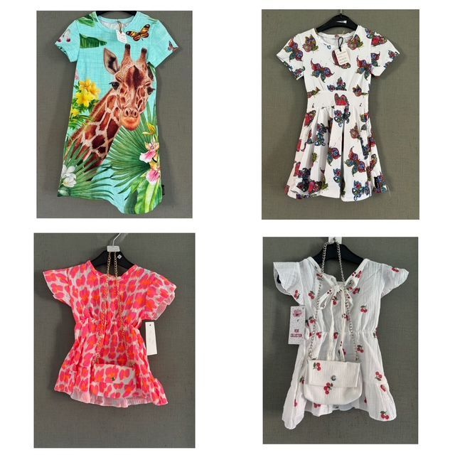 Vier kinderjurkjes hangen tegen een grijze muur: een met giraffenprint, een met bloemen/vlinders, een neonroze en een witte met een tas.
