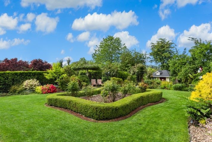 Lush green garden with neatly trimmed hedges, flowering bushes, and a small building under a bright blue sky.