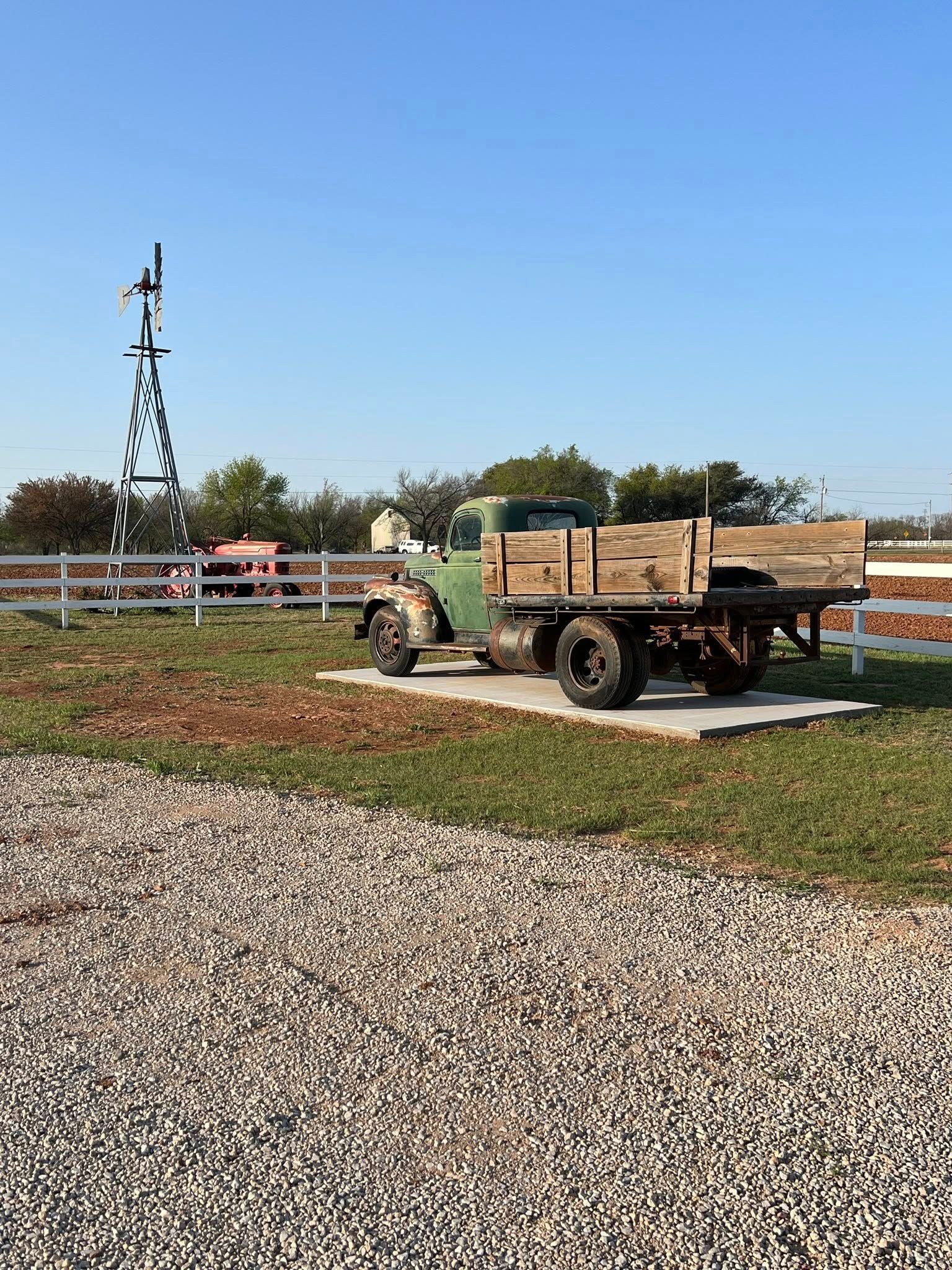 Green vintage truck on concrete slab in grassy area. Tall metal sculpture and white fence in background.