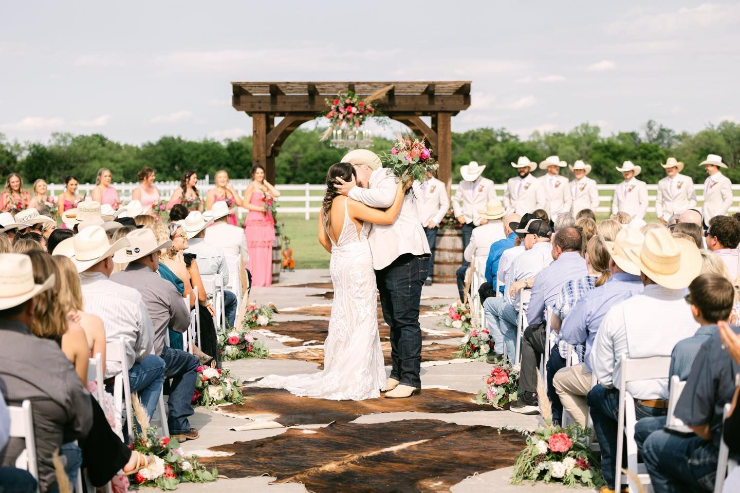 Wedding ceremony with couple embracing under an arbor, guests seated, outdoor setting.