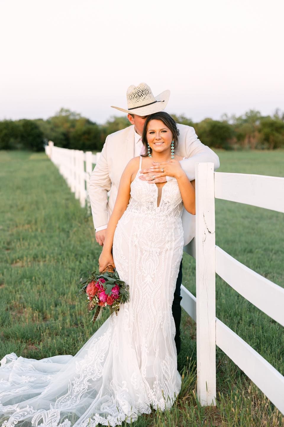 Couple at wedding. Bride in white gown leans against white fence, groom in cowboy hat stands behind her.