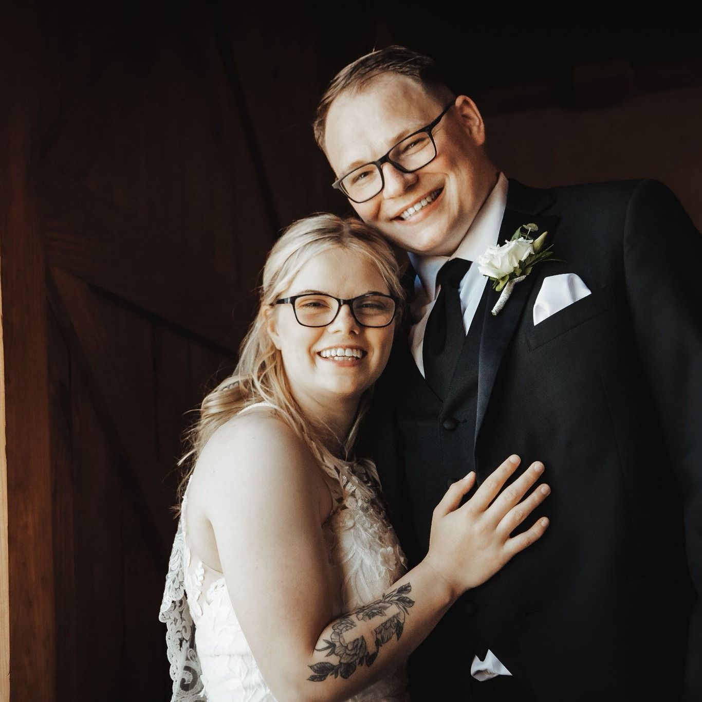 Bride and groom smiling, embracing. She wears a white dress, he wears a suit, both with glasses.