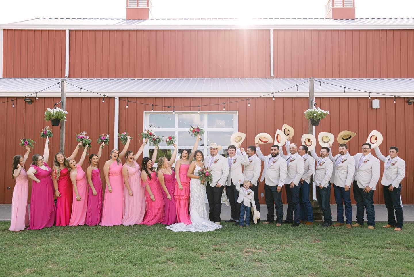 Wedding party poses in front of a red barn, women in pink dresses, men in tan shirts, cowboy hats raised.