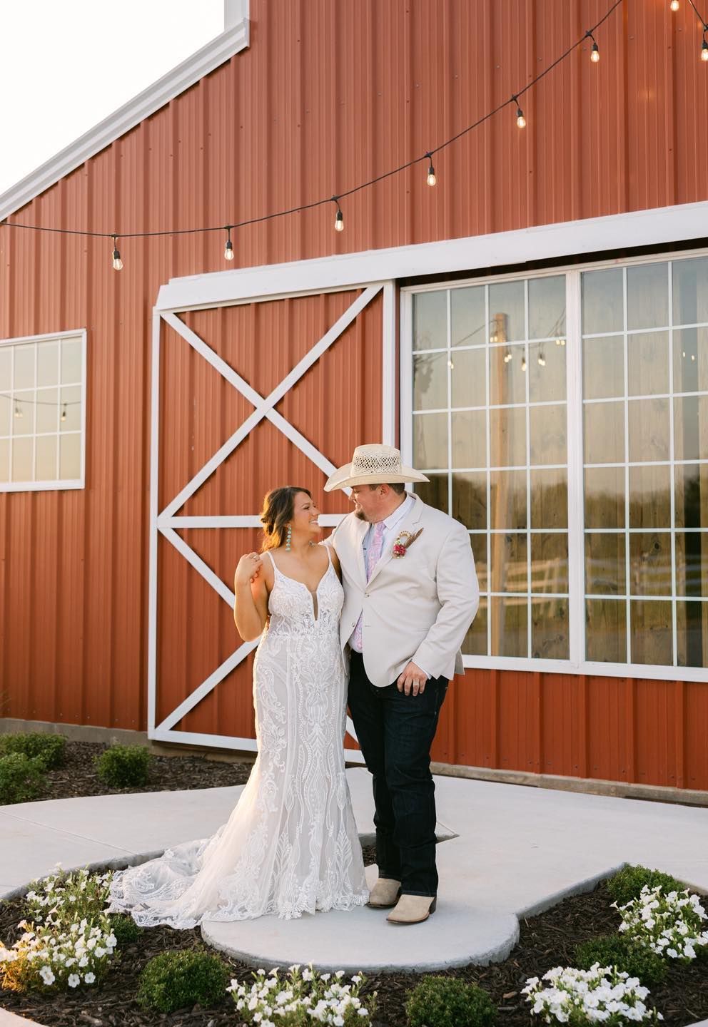 Bride and groom embrace by a red barn with string lights. She wears a white gown, he wears a cream blazer, cowboy hat.