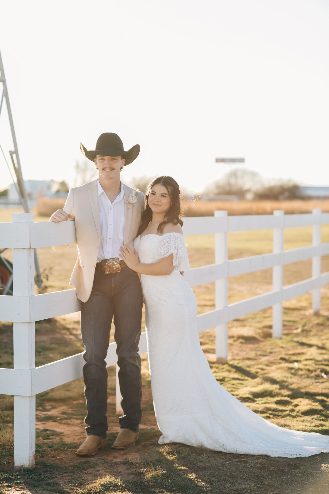 Couple in wedding attire posing by a white fence. Man in cowboy hat, beige jacket, and jeans. Woman in white off-shoulder gown.