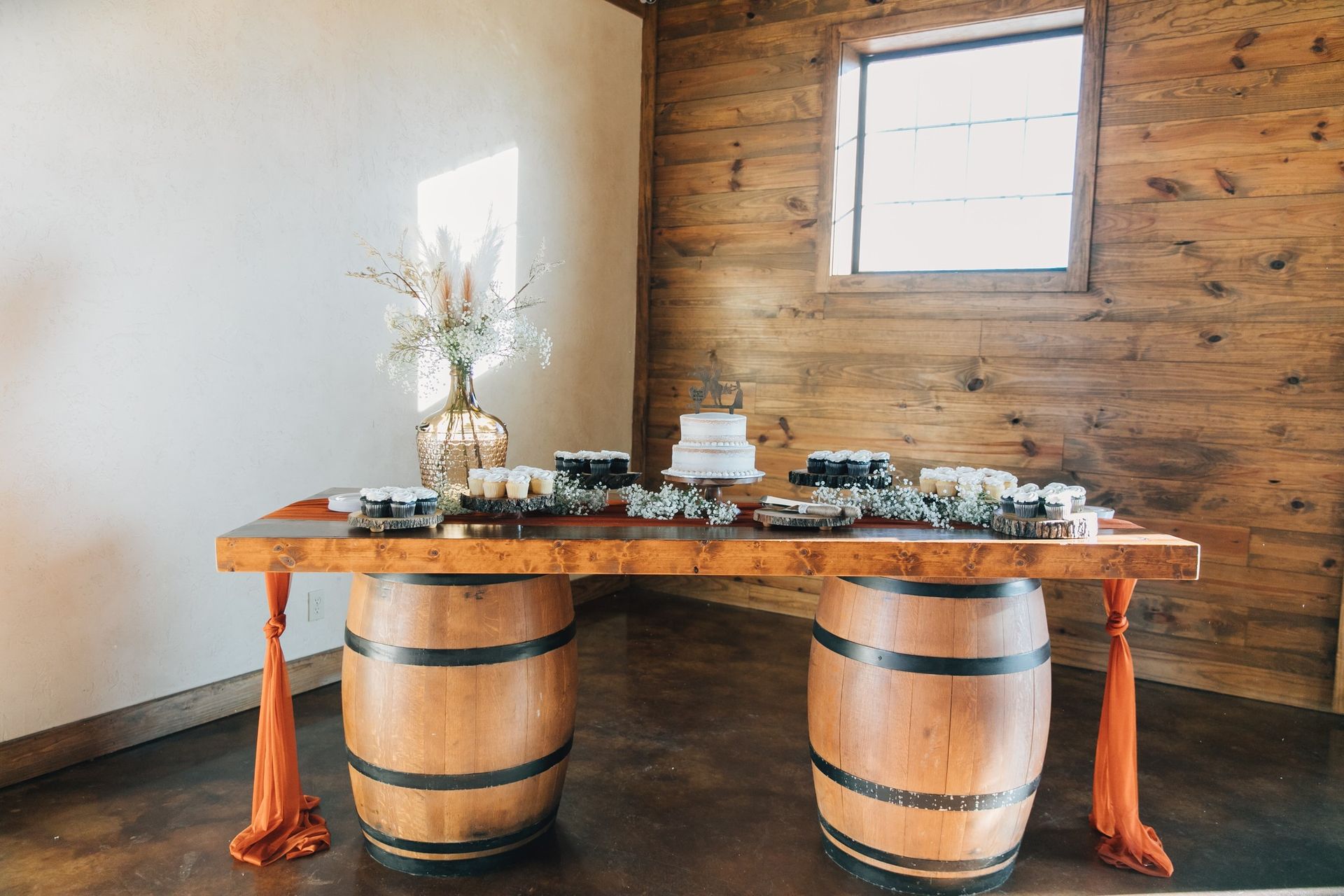 Cake table made of a wooden table top supported by two barrels. Orange fabric drapes down.