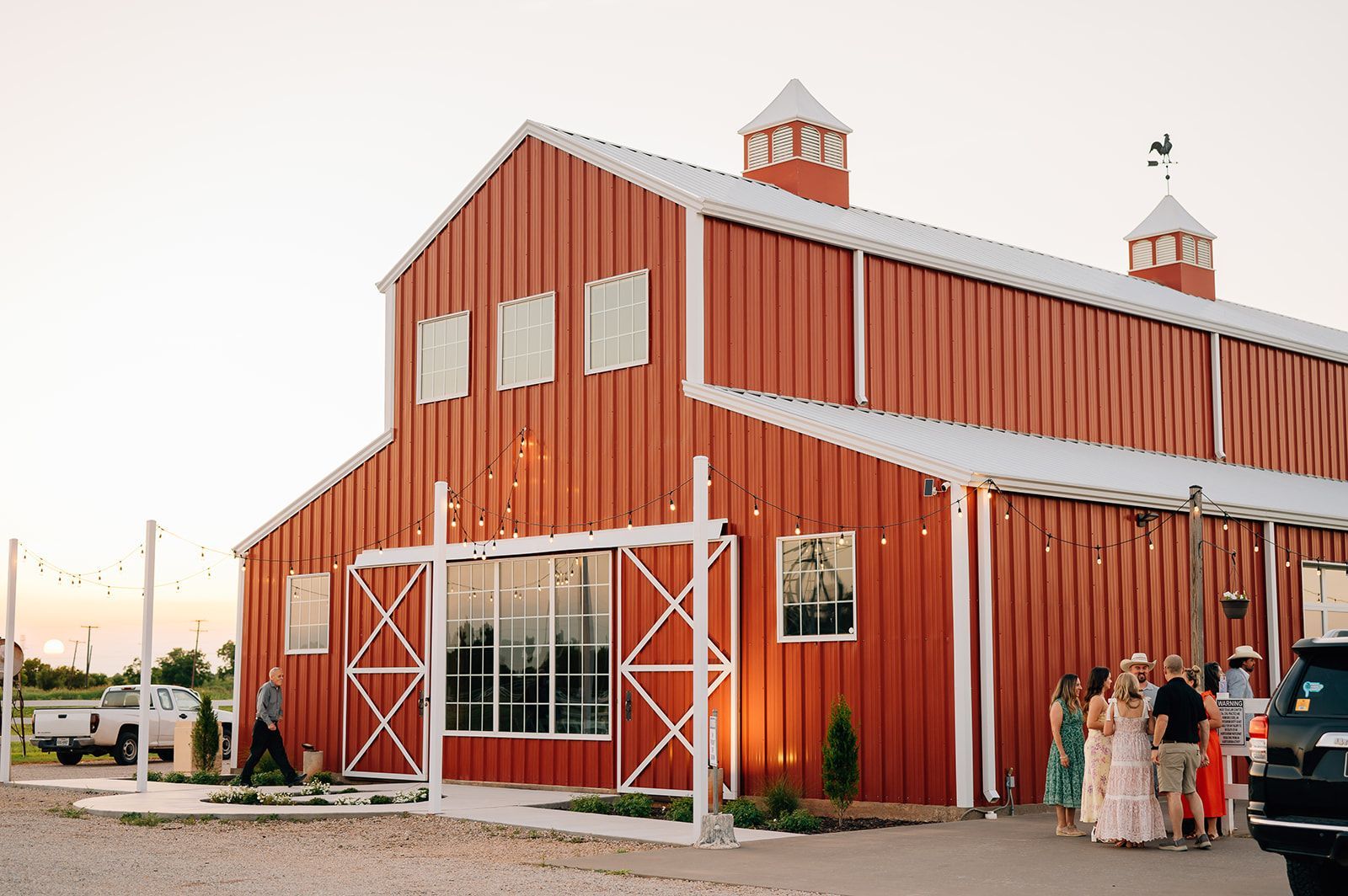 Red barn with white trim, possibly a wedding venue. People stand outside near a car.