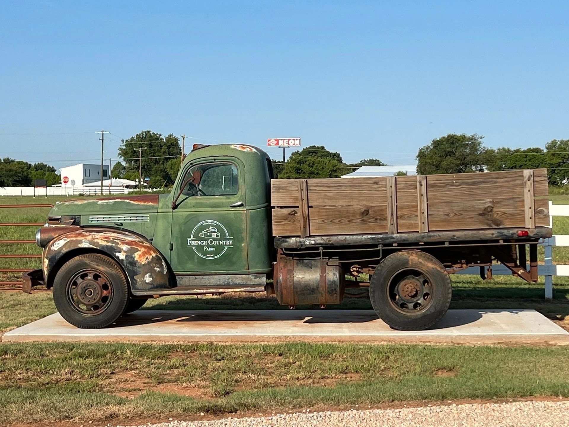 Old green pickup truck with wooden bed, parked on a concrete slab in front of a white fence under a blue sky.