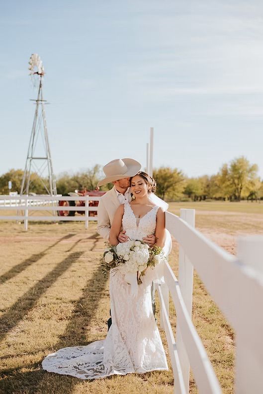 Newlyweds embrace by a white fence. Groom in cowboy hat kisses bride's head, holding her. Rustic, sunny setting.