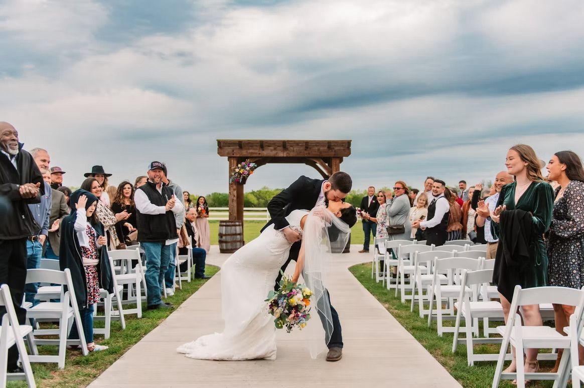 Newlyweds share a kiss after their wedding ceremony. Guests clap, outdoor setting, blue sky.