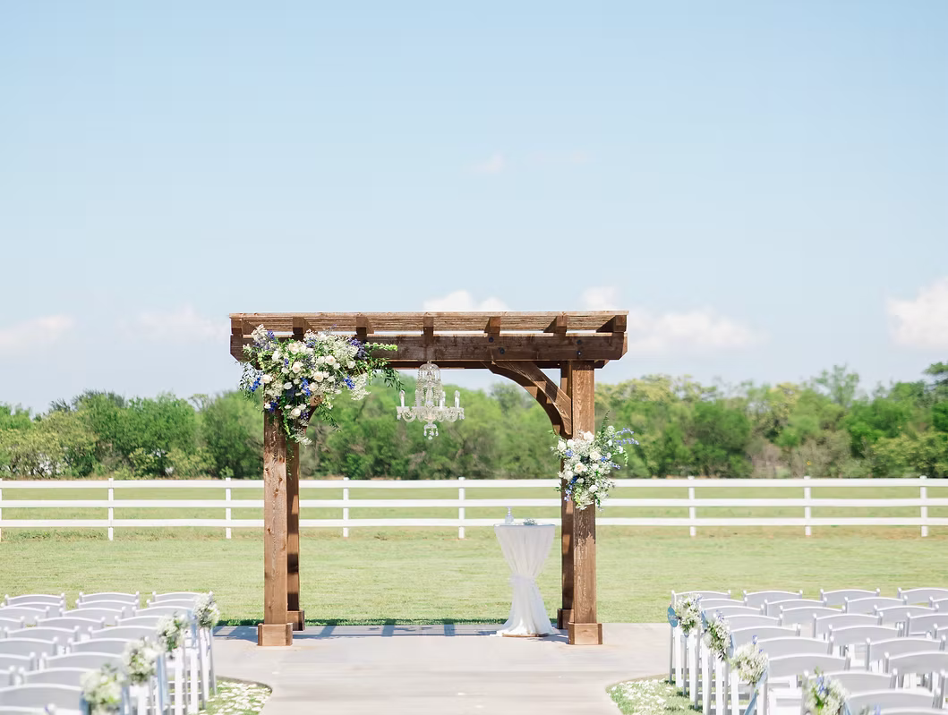 Wooden wedding arch decorated with flowers and a chandelier, set in a field with a white fence and rows of chairs.