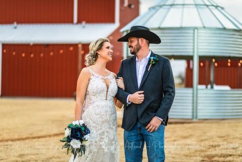 Bride and groom smiling at each other, walking outdoors near a red barn and silo gazebo.