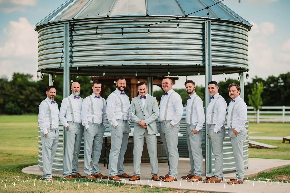 Groomsmen posing outside a metal structure. Men in light-colored suits and bow ties.