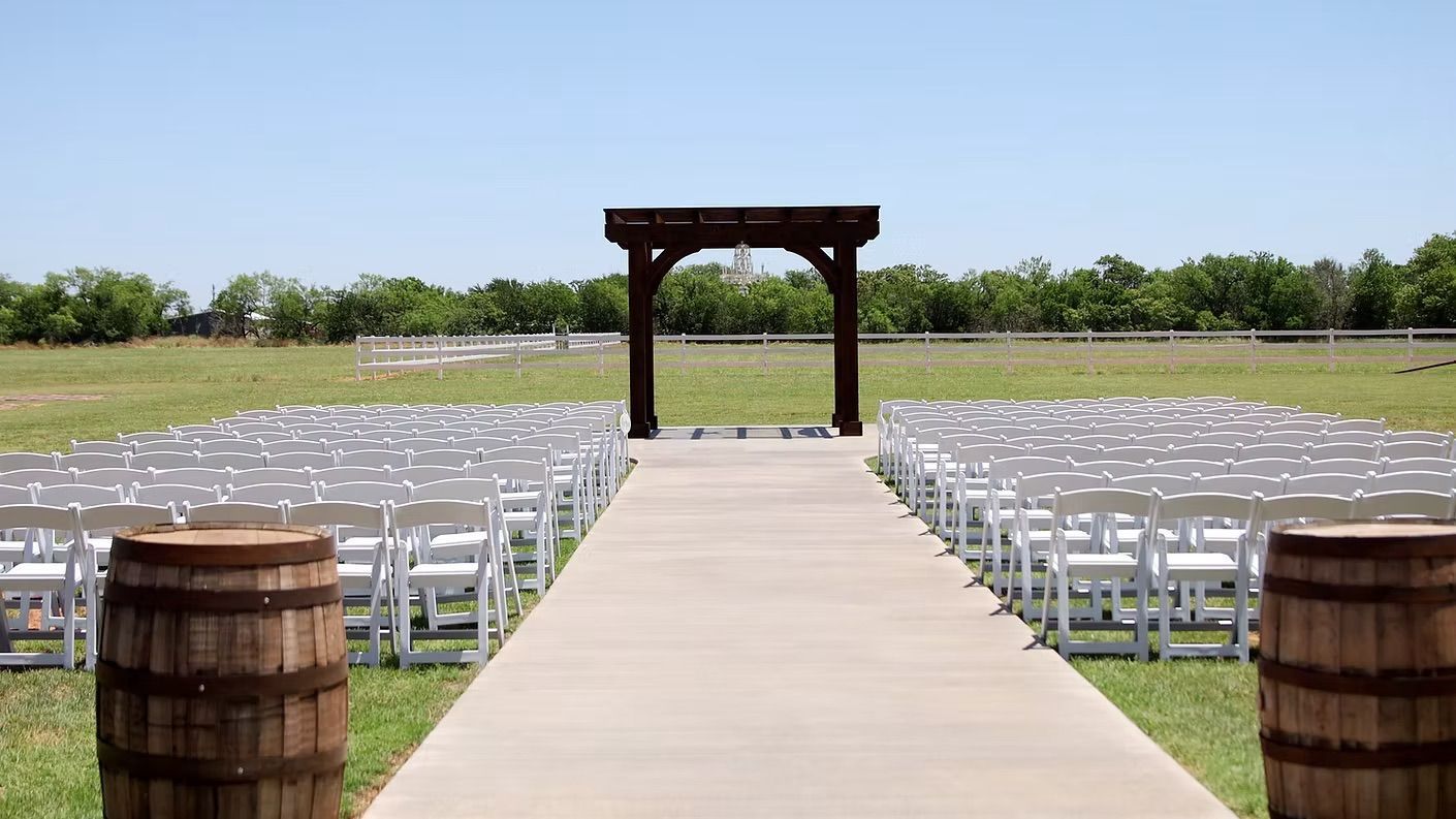 Outdoor wedding ceremony setup, rows of white chairs, aisle leading to wooden archway, barrels.