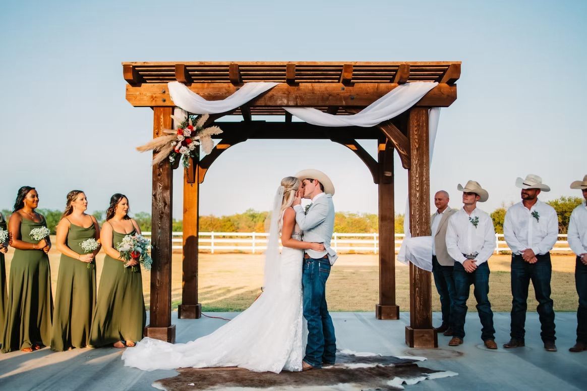Bride and groom kiss under wooden archway during outdoor wedding ceremony. Bridesmaids and groomsmen watch.
