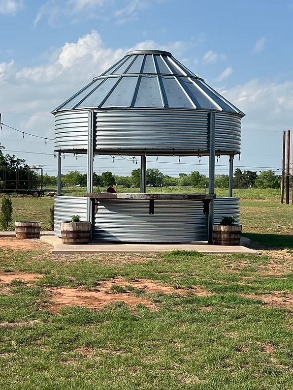 Outdoor bar made from a corrugated metal silo, featuring a wooden countertop and roof, set on concrete with barrel accents.