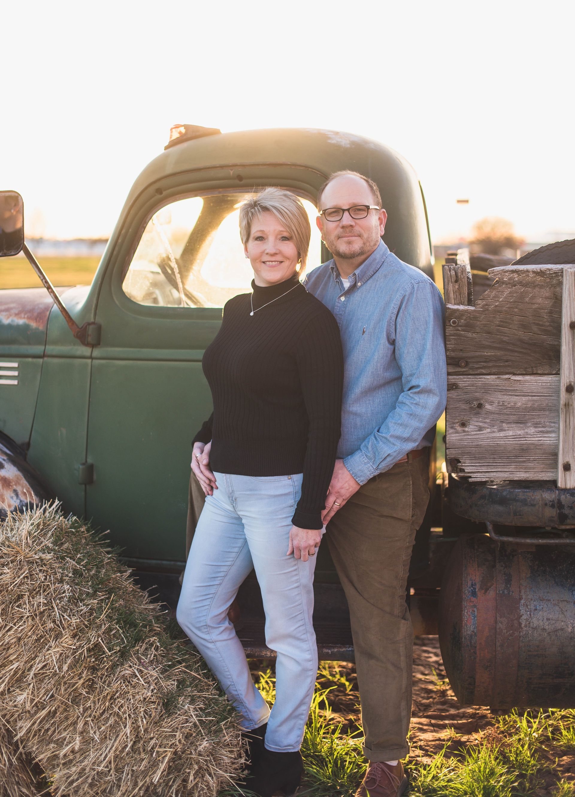 Couple poses in front of a green truck; woman in black top, jeans; man in blue shirt, glasses, brown pants.