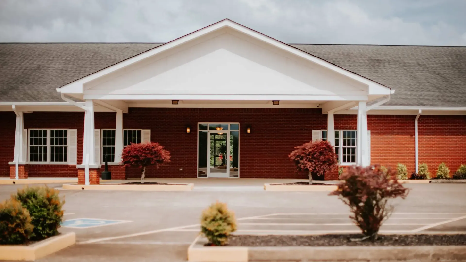 Red brick building with white trim, covered entryway, and small red bushes in front.