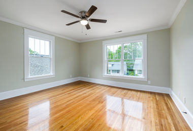 Empty room with hardwood floors, two windows, light gray walls, and a ceiling fan.