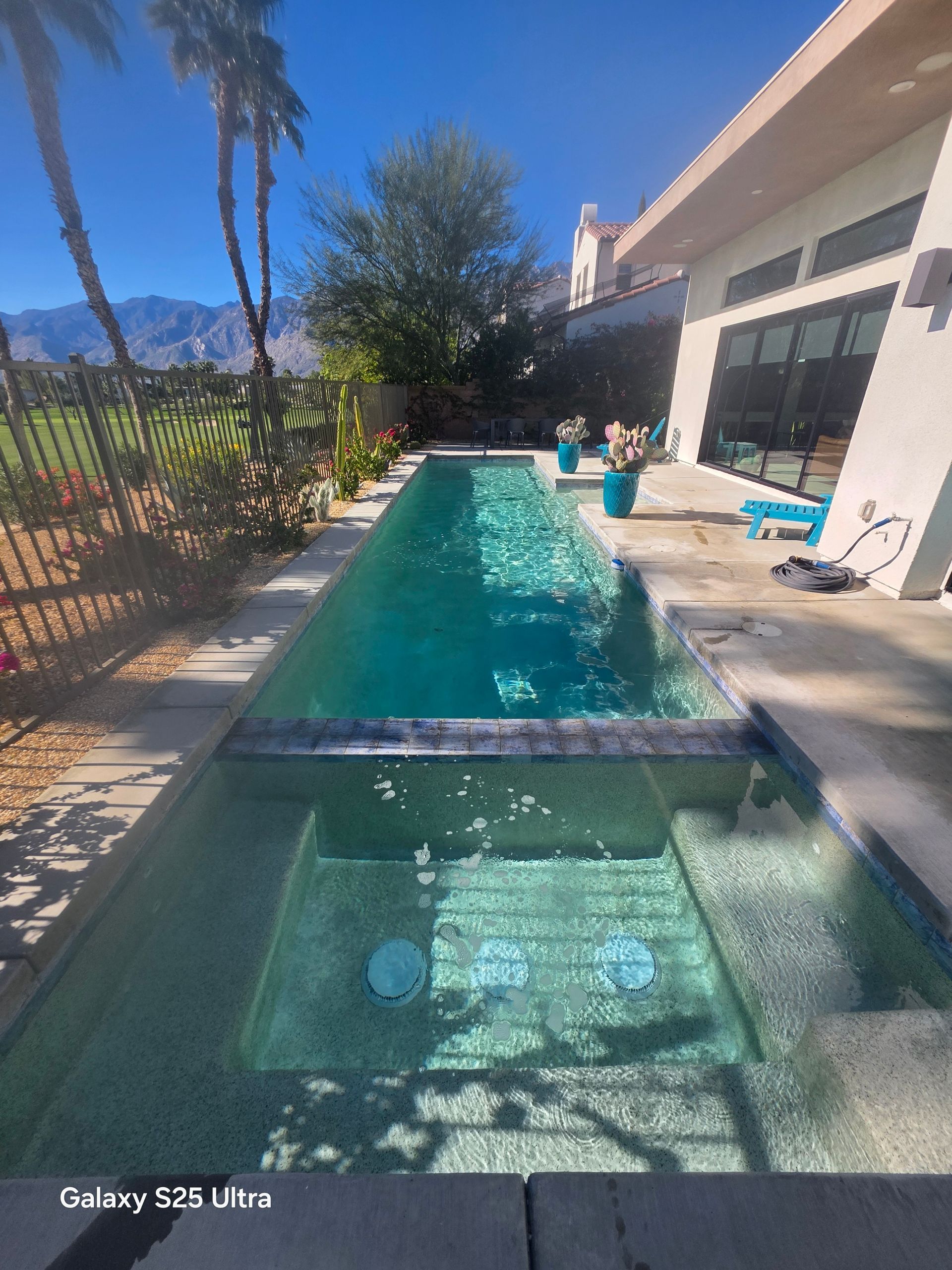 Long, narrow swimming pool with clear water, surrounded by patio and fence, under a blue sky.