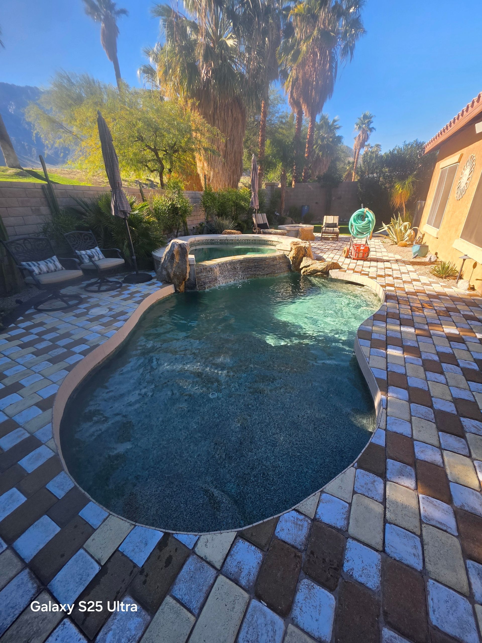 Pool with waterfall feature surrounded by brick patio, lush greenery, and palm trees. Sunny day.