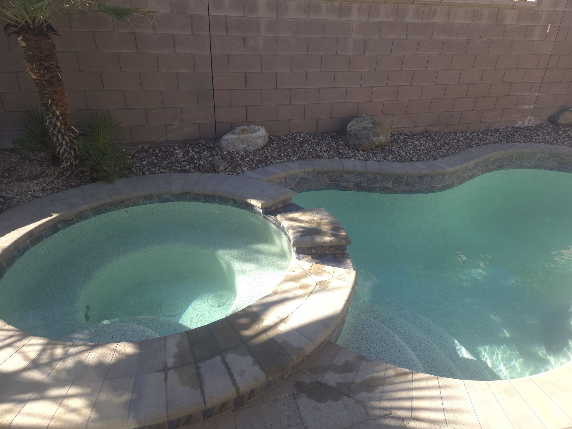 Pool and spa with light blue water, surrounded by stone and a brick wall.
