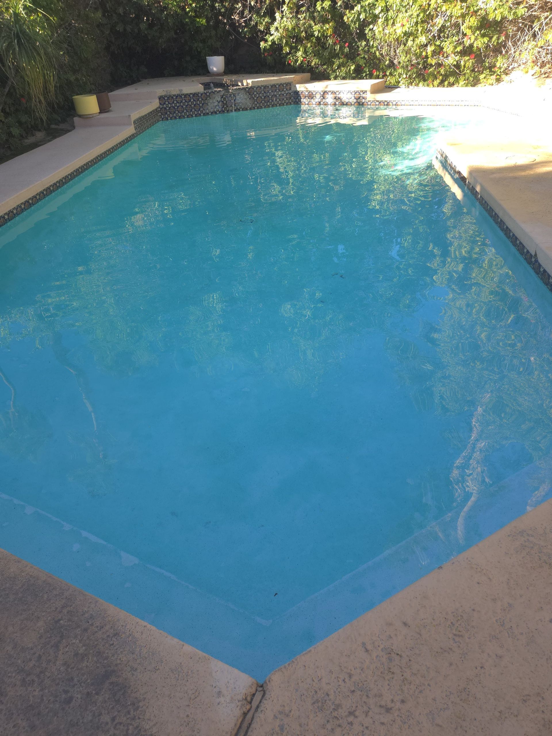 Blue swimming pool with light-colored concrete border and a small waterfall feature in a sunny outdoor setting.