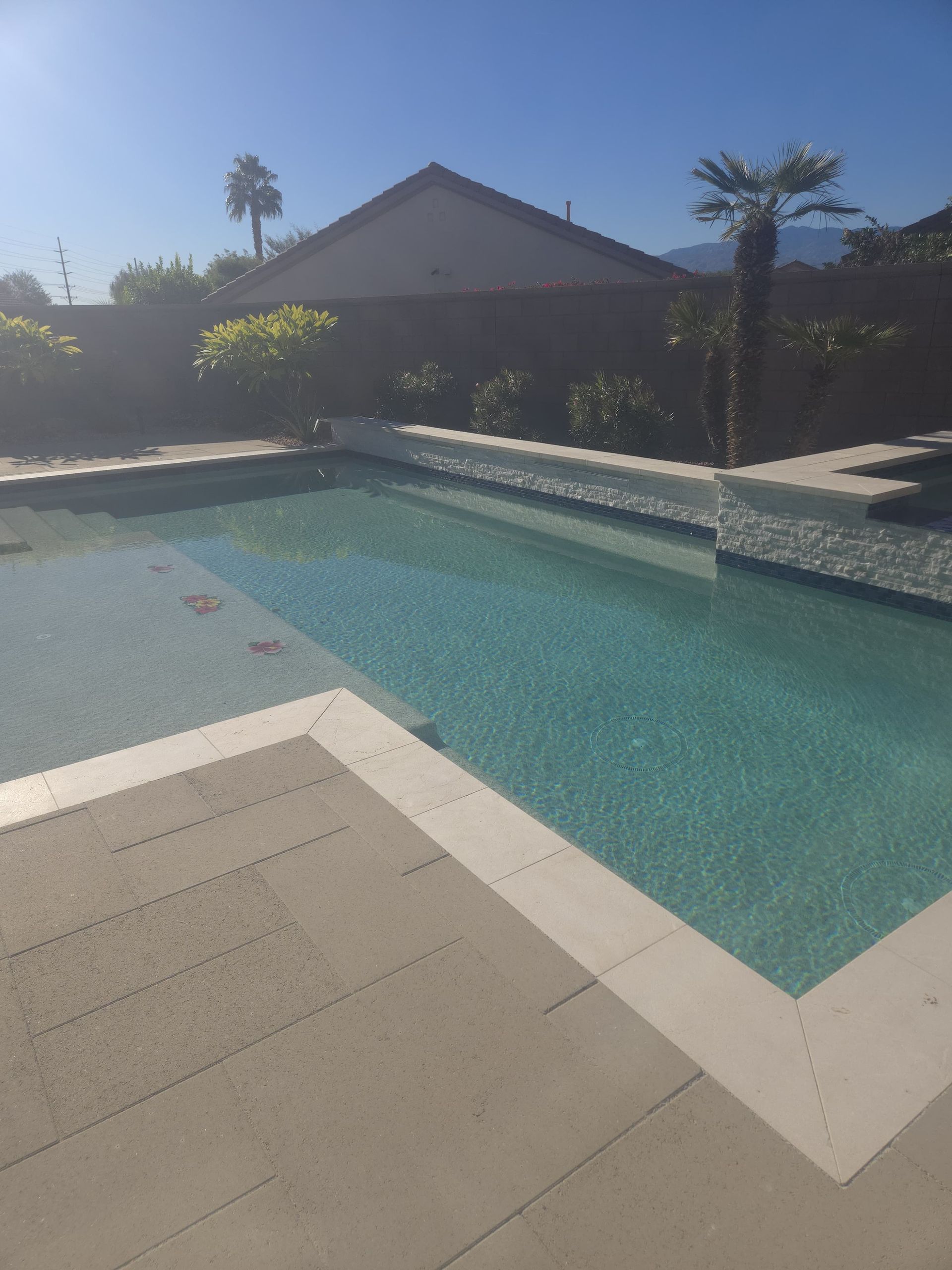 Poolside view: rectangular pool with clear water, surrounded by light-colored paving and a house in the background.
