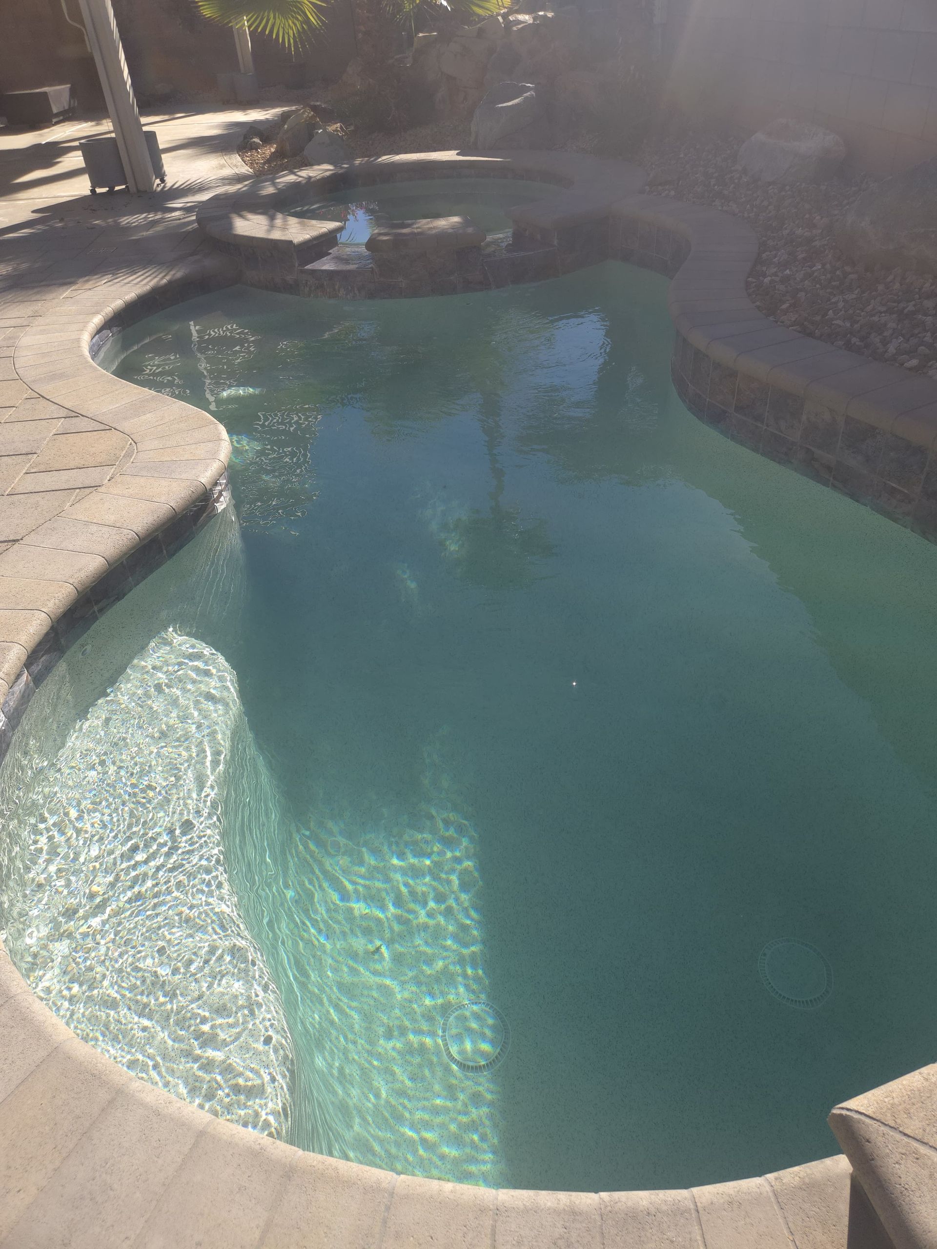 A swimming pool with clear water, surrounded by concrete and a small waterfall feature in a backyard.