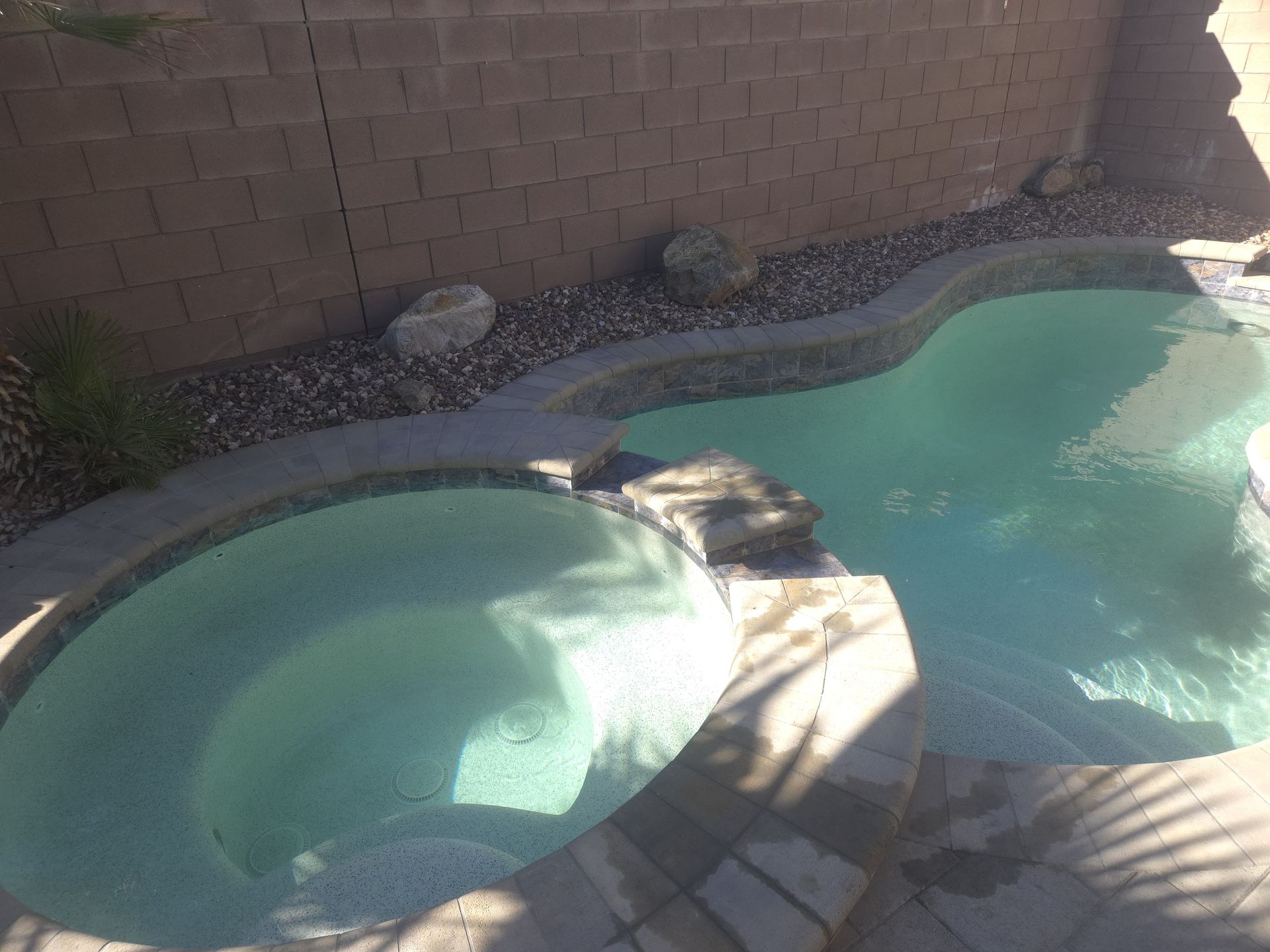 Pool with a spa, set against a brown brick wall and rocky landscape. Water is light blue.